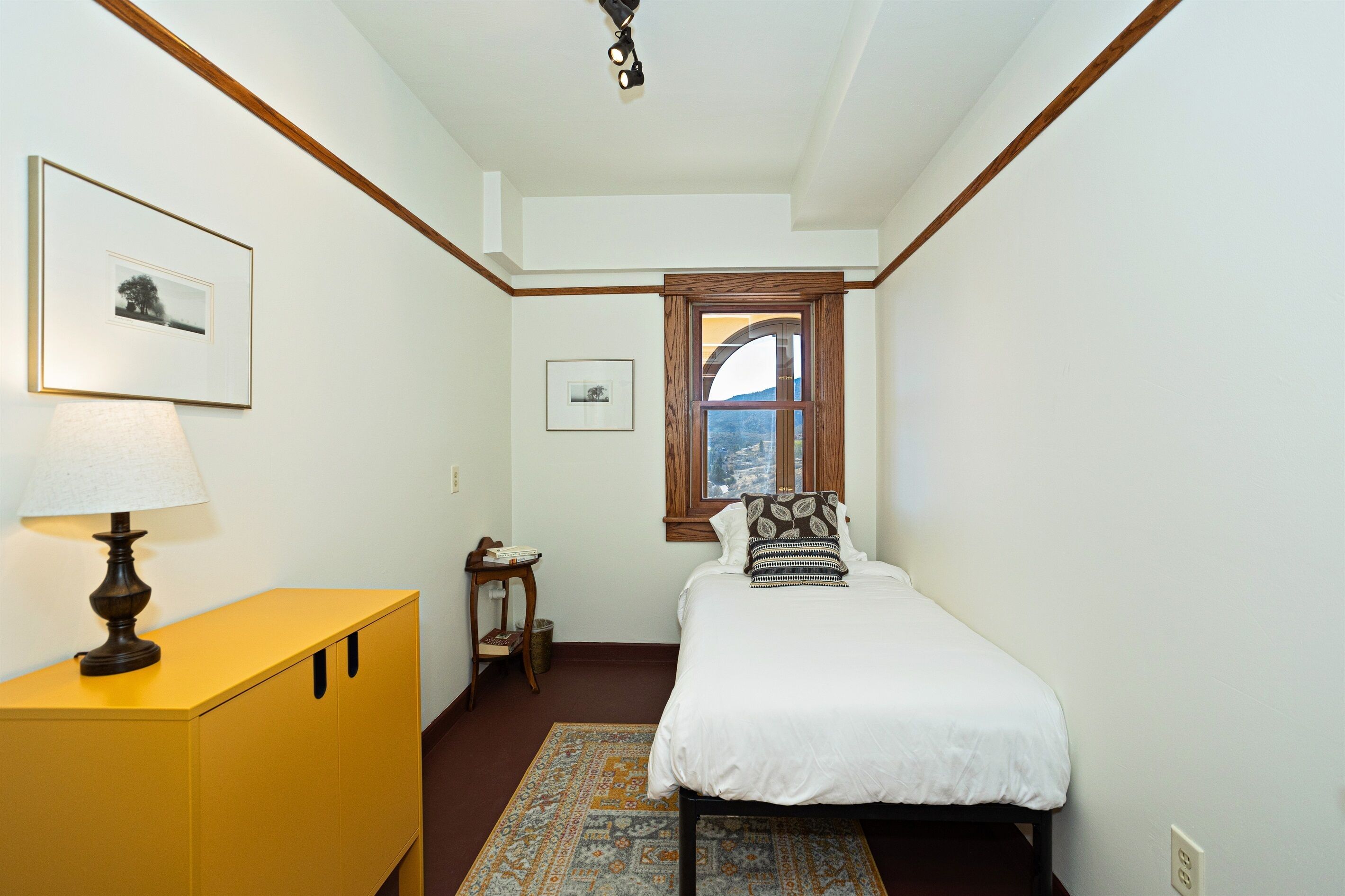 Une chambre à coucher avec un lit, une table de chevet, une lampe, un tableau encadré et une fenêtre donnant sur un paysage.
