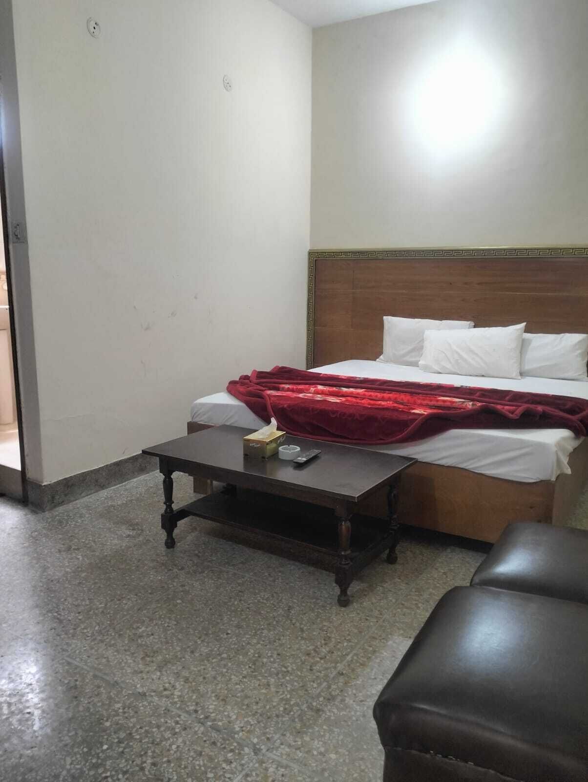 A small, simple room with a bed, a wooden nightstand, a red blanket, and a small table with a remote control and tissues.