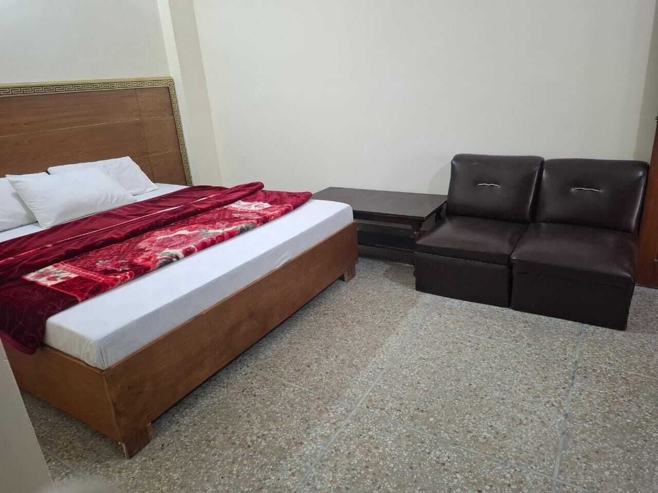 Un dormitorio con una cama, un edredón rojo, un cabecero de madera, un sofá marrón oscuro y una pequeña mesa.
