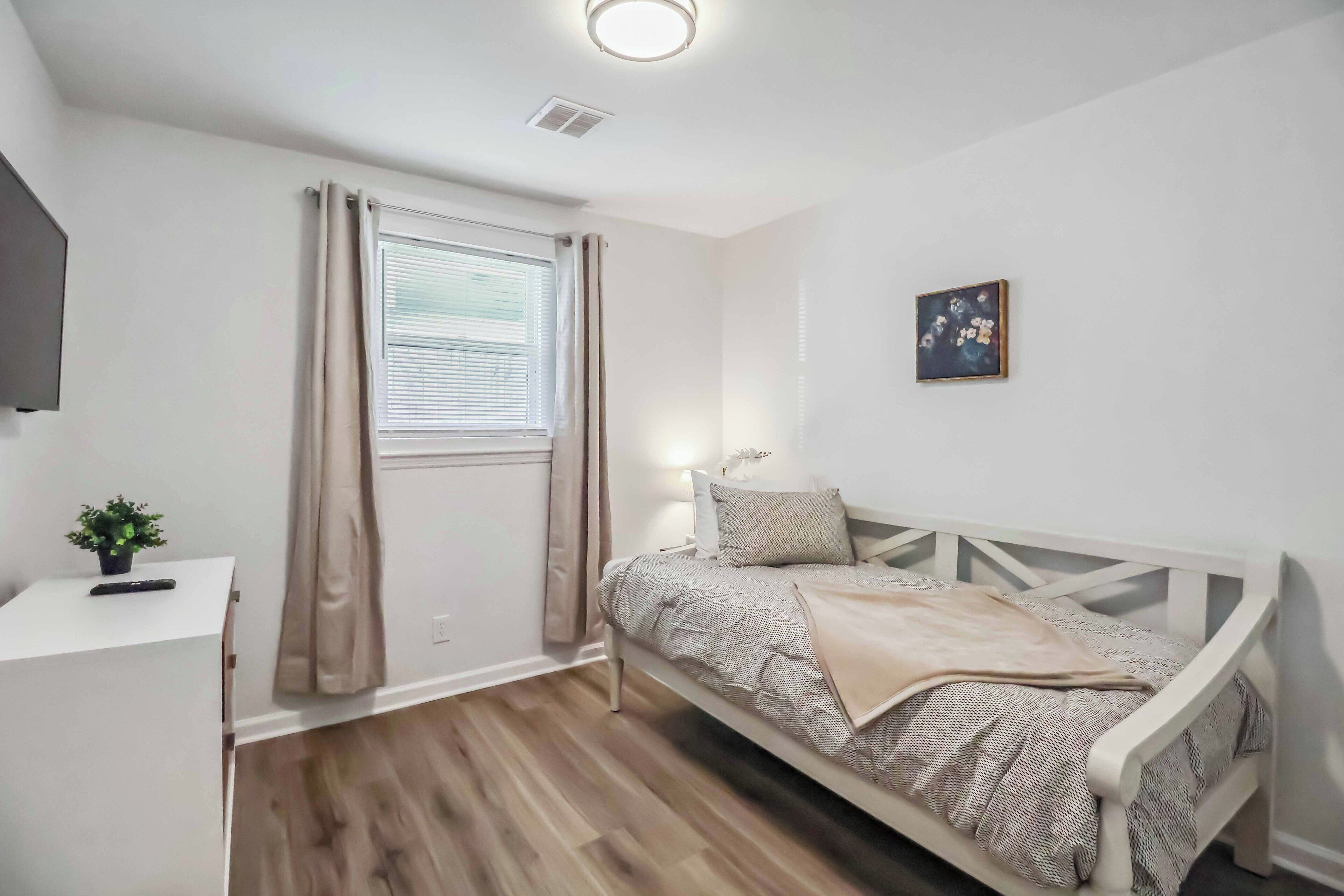 A bedroom with a bed, a desk, a plant, and a window with curtains.