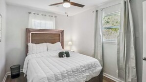 A neatly made bed with a white quilt, a wooden headboard, a bedside lamp, and a window with curtains. - Fully Furnished 2BR Near Duke & VA Hospital Ideal for Medical & Extended Stays (Durham)