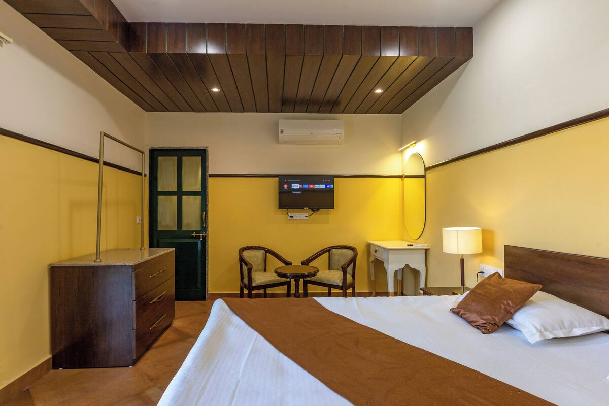 A hotel room with a bed, a desk, two chairs, a television, and an air conditioning unit.