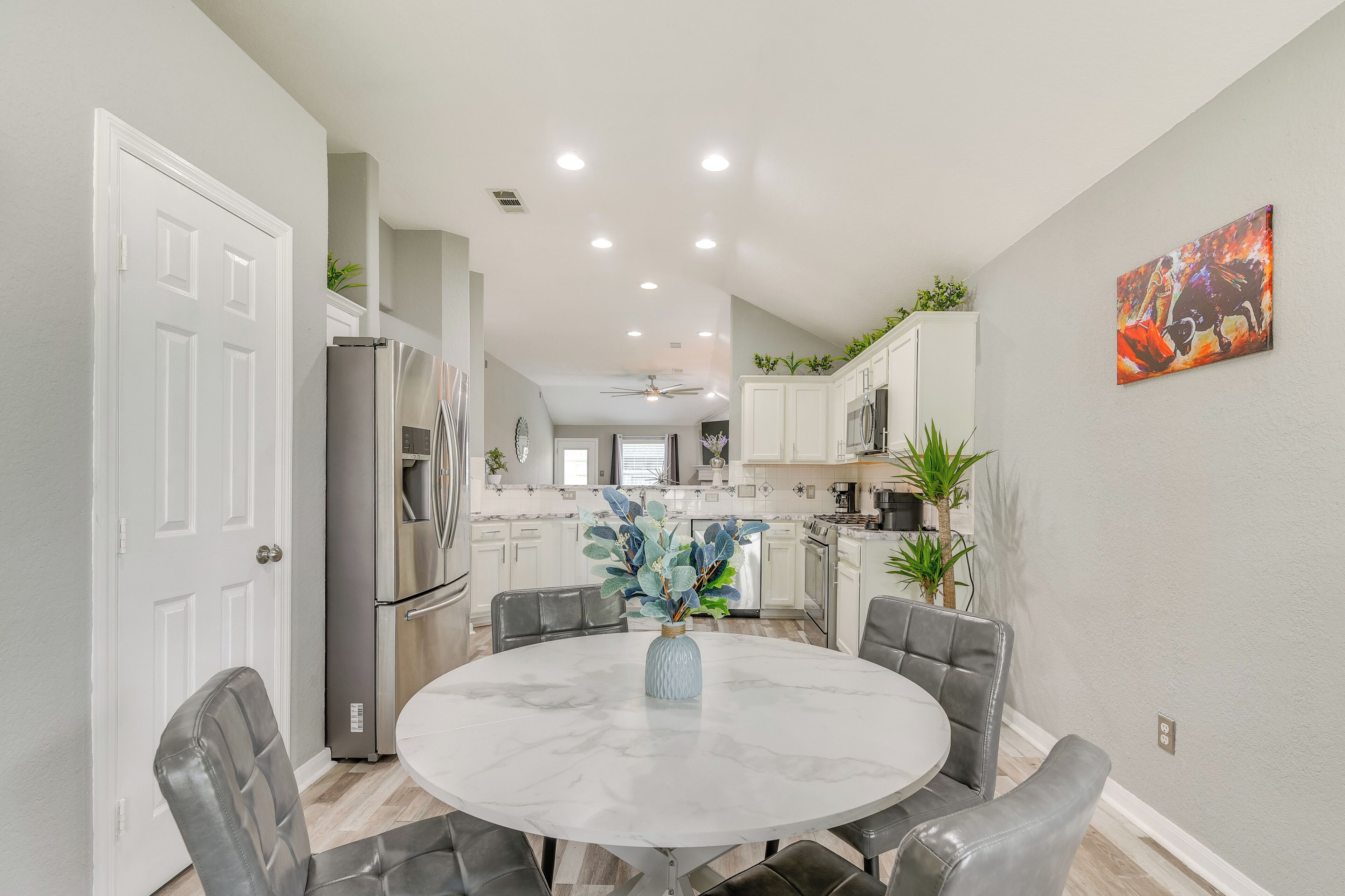 A modern kitchen with white cabinets, a stainless steel refrigerator, and a dining area with a marble table and leather chairs.
