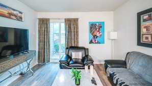 A living room with a TV, a black leather sofa, a grey sofa, a coffee table with a plant, and a painting on the wall. - 9 Mi to Dtwn Dallas: Cozy Couple's Getaway (Dallas)