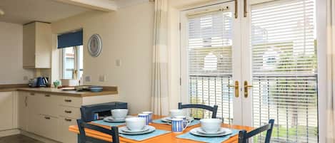 A dining area with a table set for four, chairs with cushioned seats, a kitchen in the background, and a door with blinds.