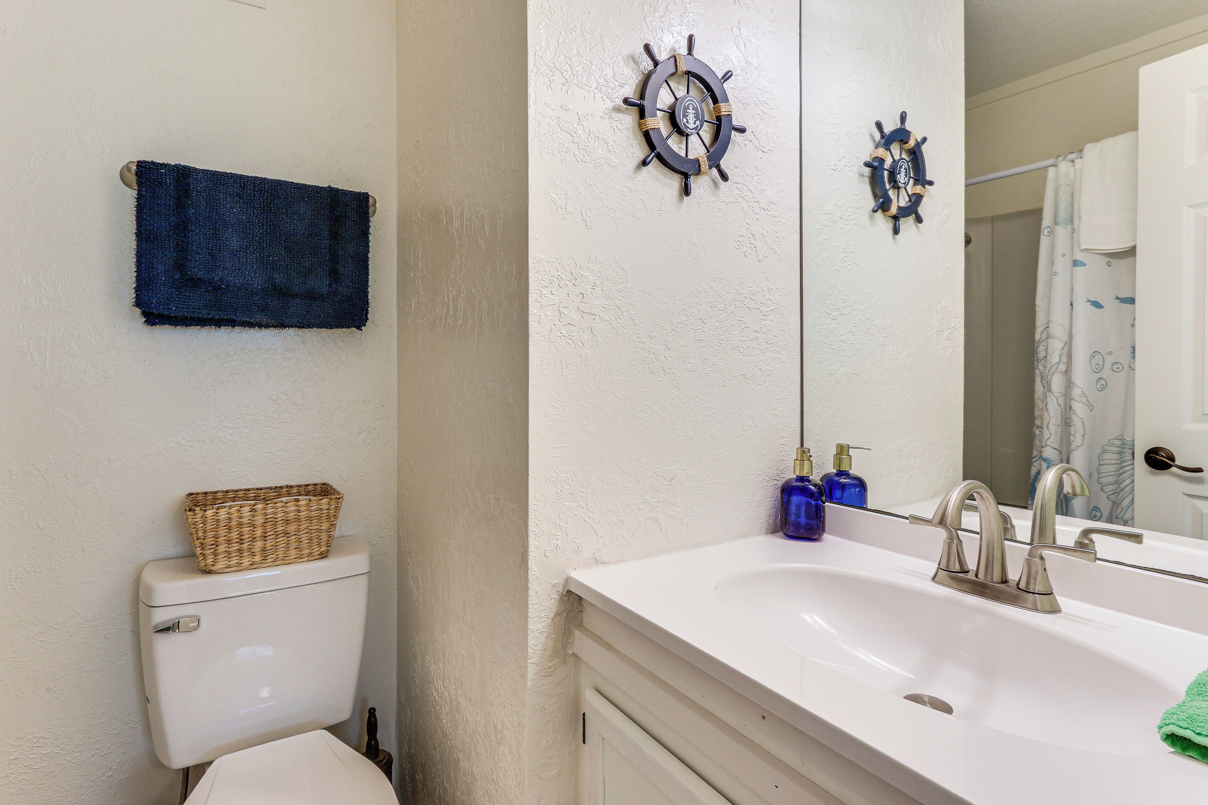 A bathroom with a white sink, a ship's wheel decoration, a blue towel, and a wicker basket on the toilet.