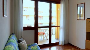 Une pièce avec un plancher en bois, un lit recouvert d’une courtepointe aux couleurs vives, une chaise en bois, un meuble en bois et une porte-fenêtre coulissante donnant sur un balcon où se trouvent une chaise blanche et une table.