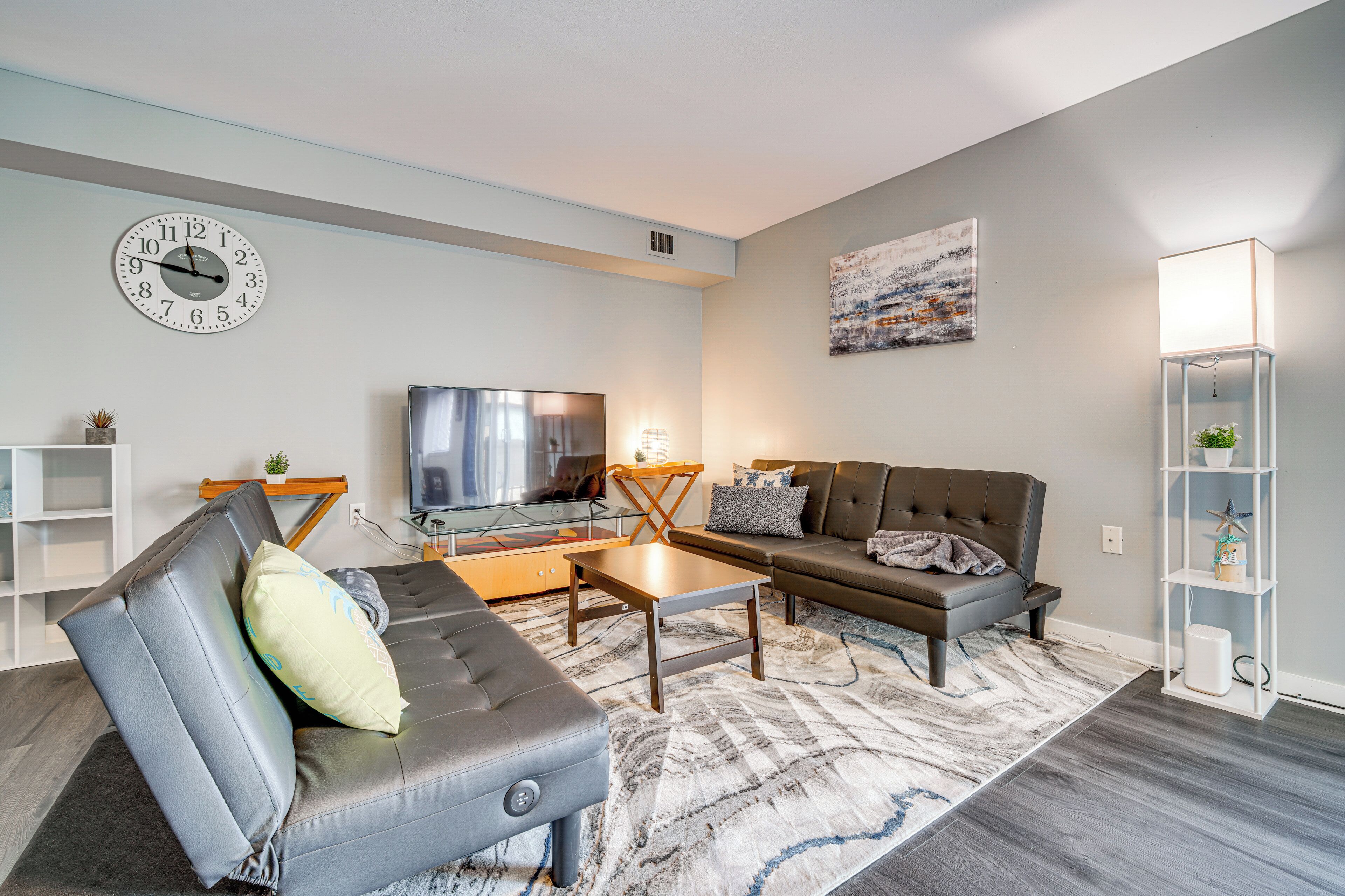 A modern living room with a sofa, coffee table, and a TV. There is a clock on the wall and a painting above the sofa.