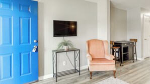 Une pièce avec une porte bleue, un téléviseur fixé au mur, une chaise et une petite table sur laquelle se trouve une plante.
