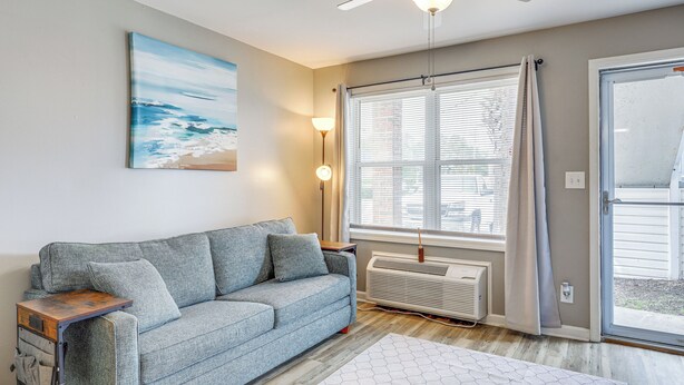 Un salon avec un canapé gris, un ventilateur de plafond, un tableau représentant une plage et une grande fenêtre avec des rideaux.