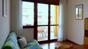 A room with a wooden floor, a bed with a blue and green striped bedspread, a wooden chair, a wooden cabinet, and a sliding glass door leading to an outdoor area with a white chair and a table. - Calm and cozy stay in Bibione (Bibione)