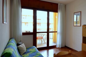 A room with a wooden floor, a bed with a blue and green striped bedspread, a wooden chair, a wooden cabinet, and a sliding glass door leading to an outdoor area with a white chair and a table. - Calm and cozy stay in Bibione (Bibione)