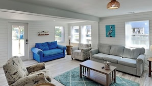 Un salón amplio con un sofá azul, un sofá gris y sillones estampados. Hay una mesa de centro y una cesta de mimbre en el suelo.