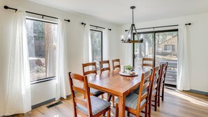 A dining room with a wooden table and chairs, large windows with curtains, and a hanging light fixture.
