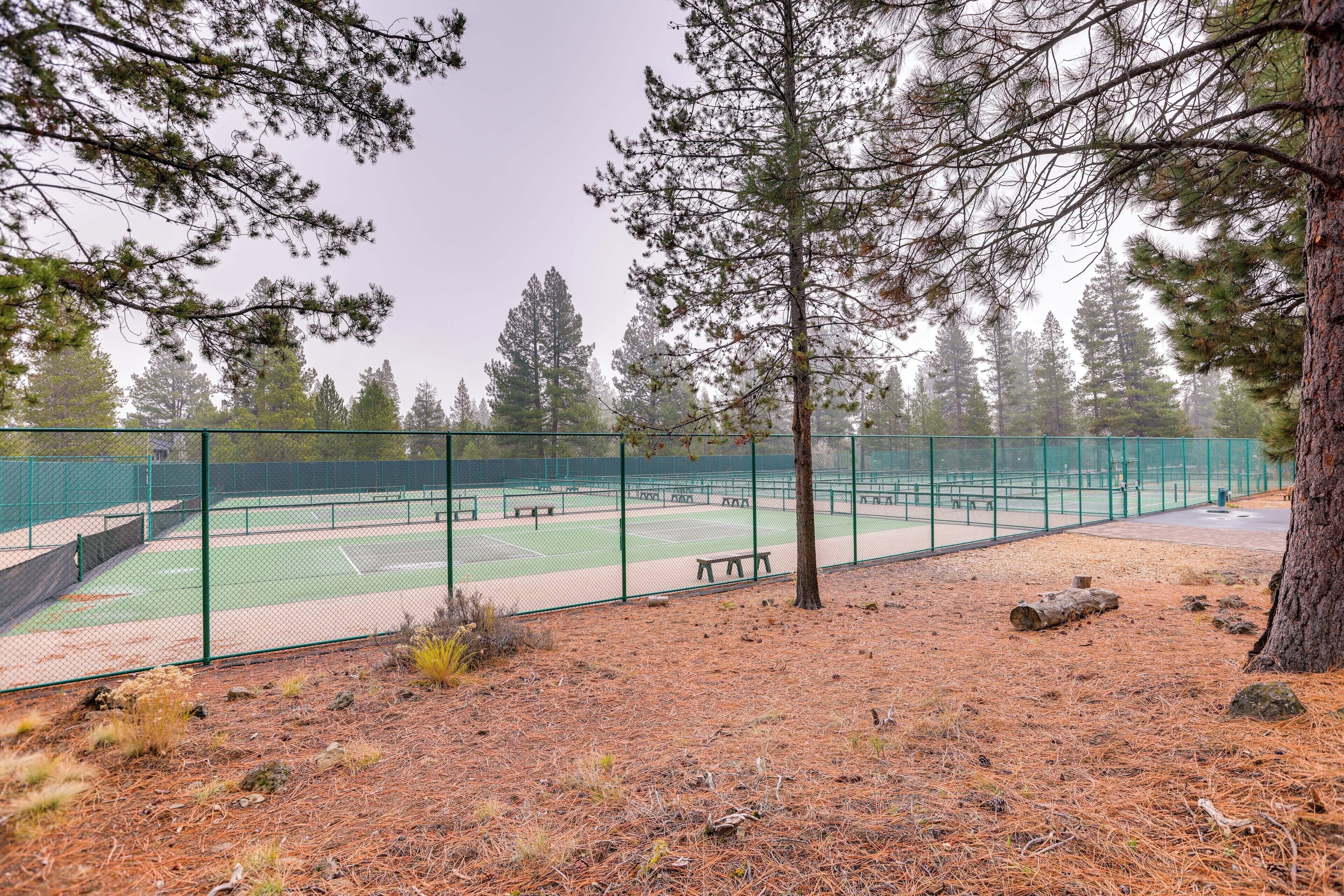 Un court de tennis entouré d’une clôture verte, avec des bancs et des arbres en arrière-plan.