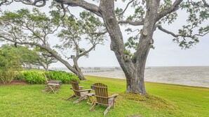 Un grand arbre sous lequel se trouvent un banc et des chaises, une zone herbeuse et, au fond, une étendue d’eau.