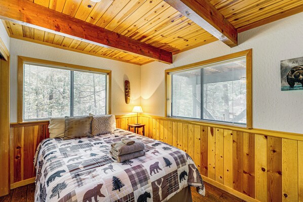 Un dormitorio acogedor con techo de madera, ventanas amplias con persianas, una cama con colcha y una lámpara en la mesita de noche.