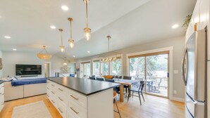 A modern kitchen with a large island, white cabinetry, and stainless steel appliances. There are pendant lights, a dining area with a table and chairs, and large windows providing natural light. - Peaceful Cabin w/ Dock on Lake Pend Oreille (Sagle)