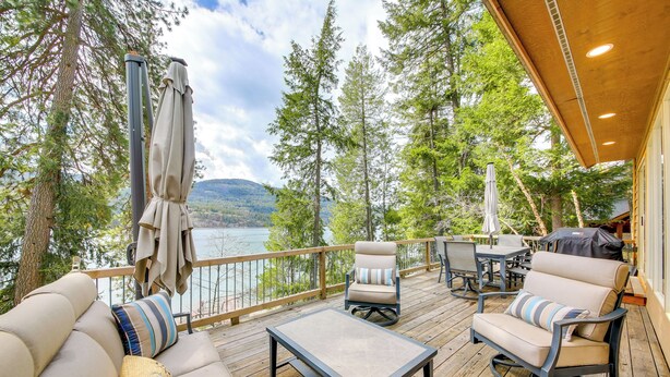 A wooden deck with outdoor furniture, including a sofa, chairs, and a table, overlooking a lake and surrounded by trees. - Peaceful Cabin w/ Dock on Lake Pend Oreille (Sagle)