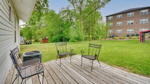 A wooden deck with two metal chairs and a grill, overlooking a grassy yard with trees and a brick building in the background. - 2 Mi to Downtown: Greensboro Home By Park! (Greensboro)