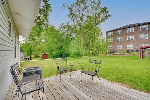 A wooden deck with two metal chairs and a grill, overlooking a grassy yard with trees and a brick building in the background.