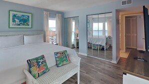 Une chambre avec un grand lit, un banc blanc agrémenté de coussins colorés, un tableau au mur et une porte-fenêtre coulissante donnant sur un balcon.