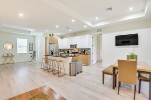 Una cocina moderna con una isla central, taburetes de bar, una mesa de comedor con sillas y un televisor de pantalla plana montado en la pared.