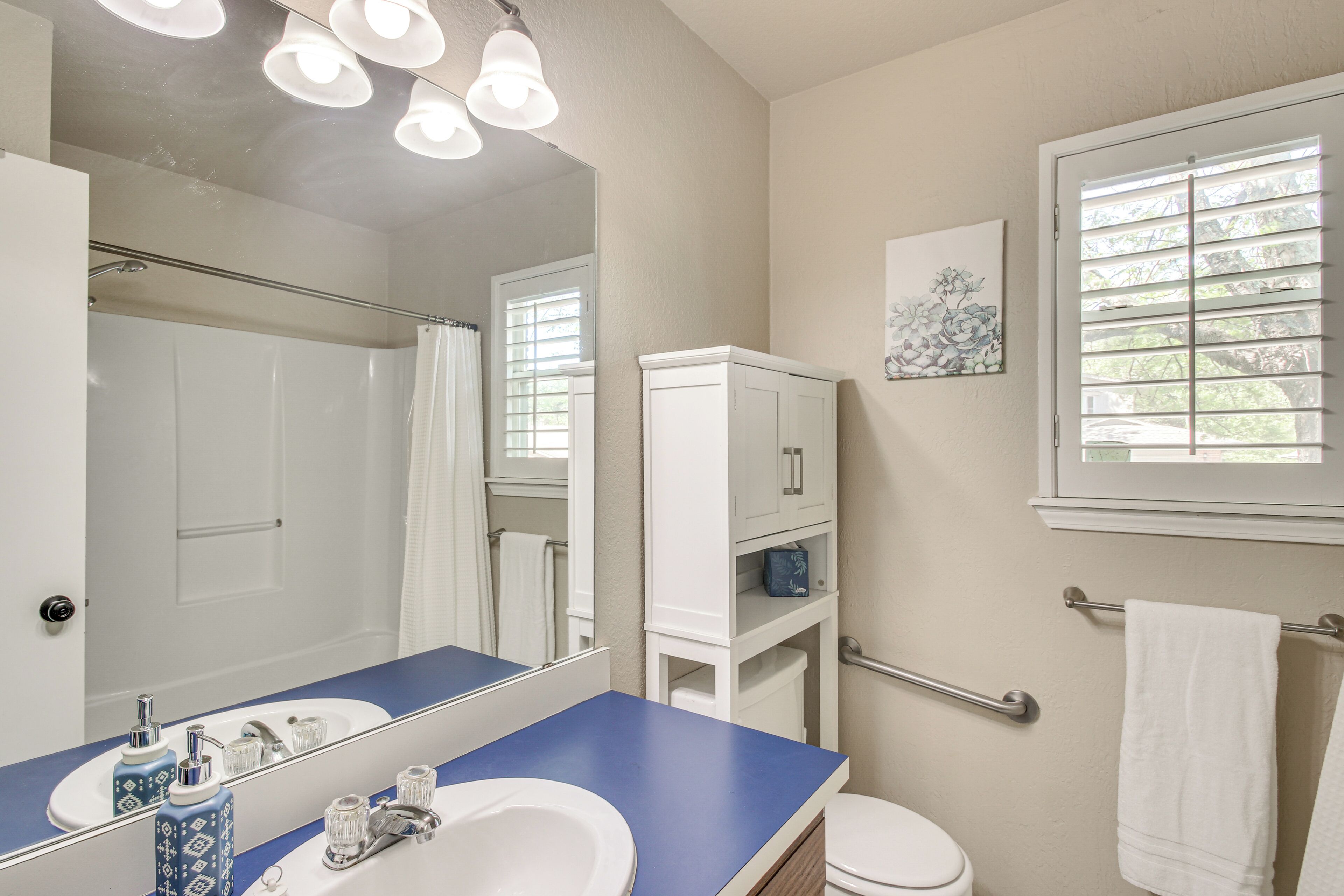 A bathroom with a blue countertop, a white sink, a mirror, a white toilet, a window with blinds, and a towel rack.