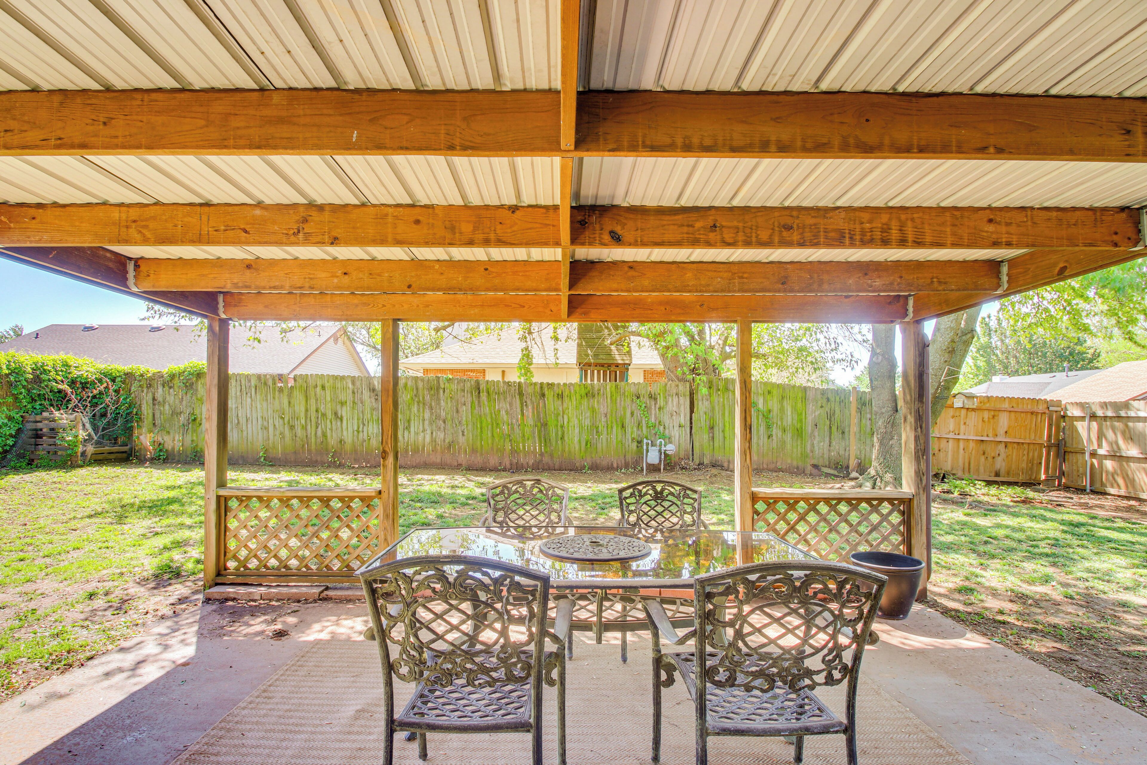 Un patio techado con una mesa y sillas, techo de madera, y vista a un jardín trasero con cerco y árboles.