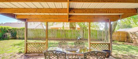 Un patio techado con una mesa y sillas, techo de madera, y vista a un jardín trasero con cerco y árboles.