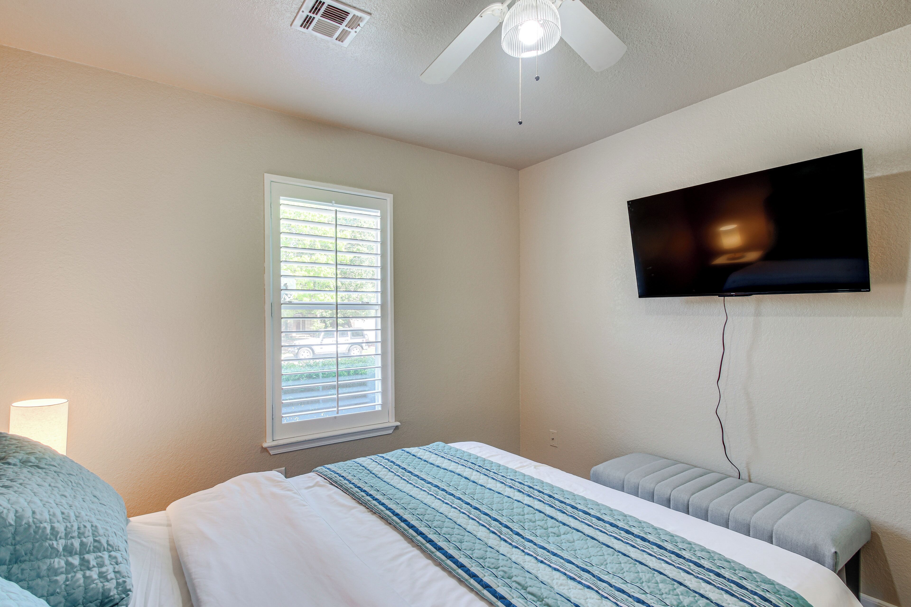 A bedroom with a bed, a window, a ceiling fan, and a TV mounted on the wall.