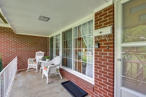 A covered porch with wicker furniture, a brick wall, and a large window with curtains. - Mid-century 1950s Home W/ Carport Near Route 66 (Monett)