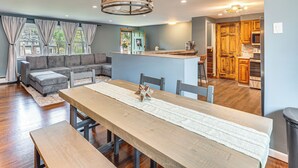 Un vaste espace de vie comprenant une grande table à manger en bois, un canapé gris et une cuisine en arrière-plan.