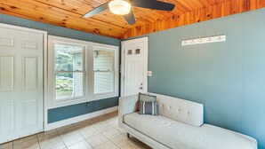 Une pièce avec un plafond en bois, un ventilateur de plafond, un luminaire, une porte blanche, une fenêtre, un mur bleu clair, un canapé beige avec des coussins et un sol carrelé.