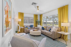 Ett modernt vardagsrum med en beige soffa, en rund pall och ett litet sidobord. Det finns en takfläkt, gula gardiner och ett stort fönster med persienner.