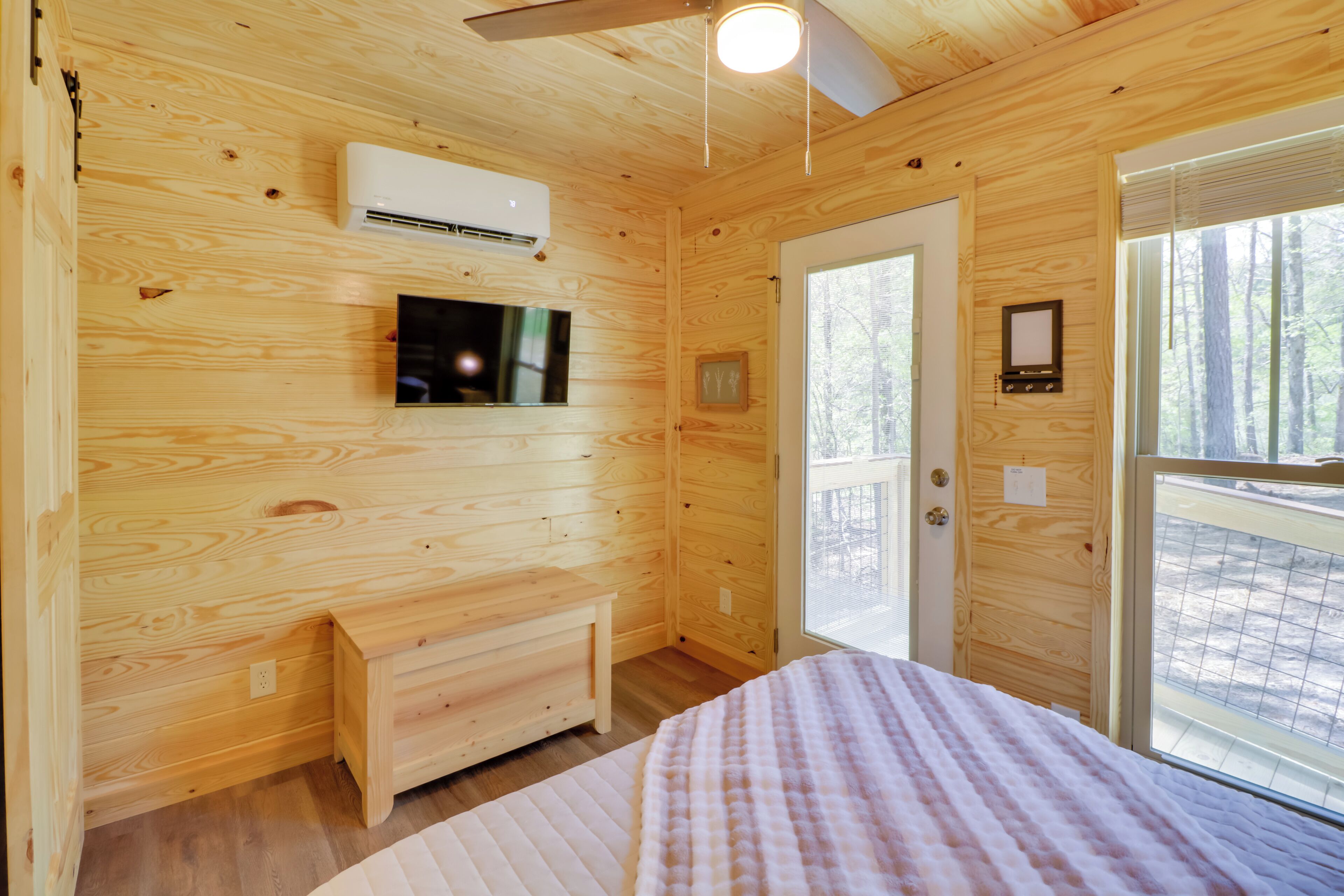 A wooden cabin bedroom with a bed, a TV, an air conditioning unit, and a door leading outside.