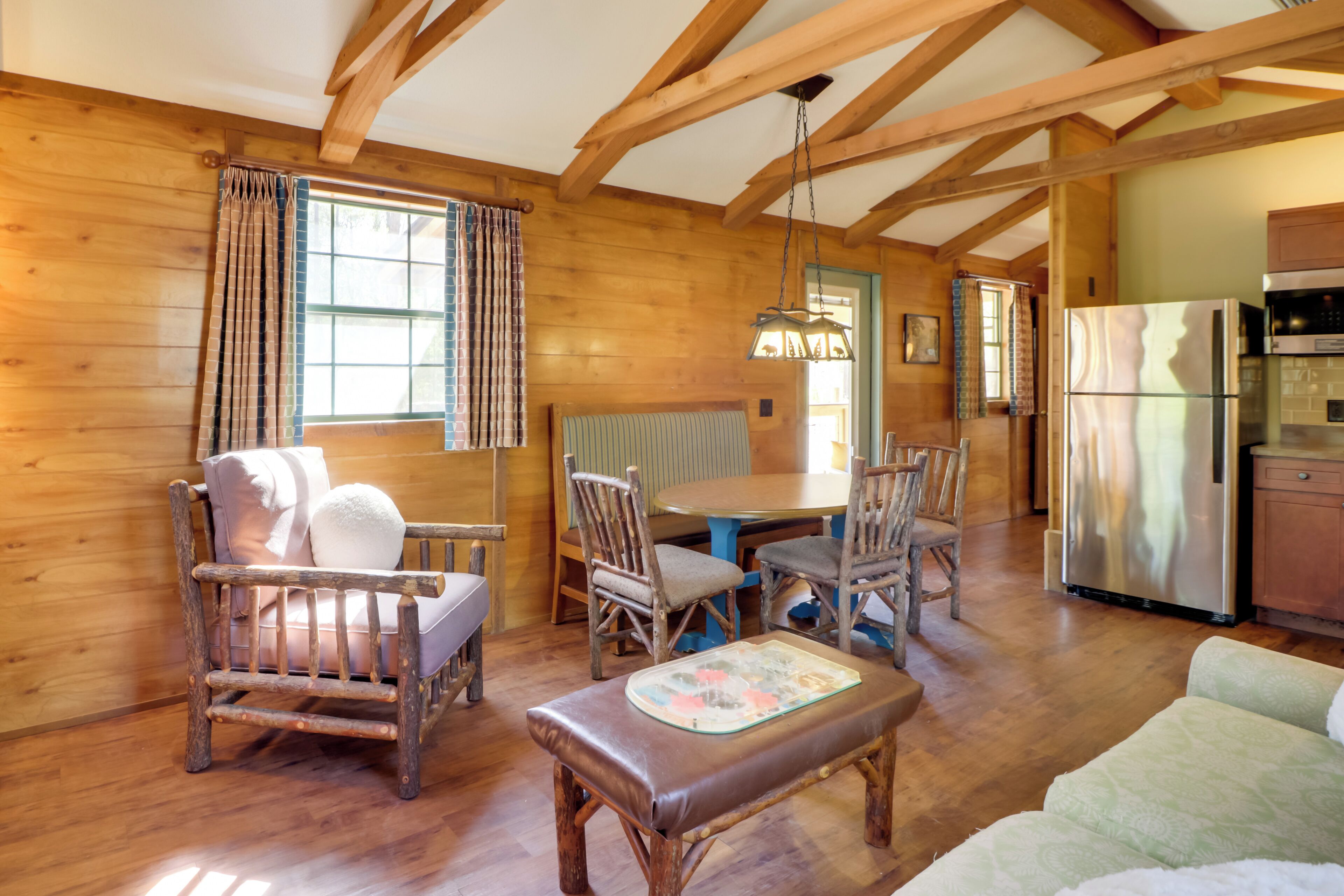 A cozy wooden cabin interior with a dining table, chairs, a sofa, and a refrigerator.