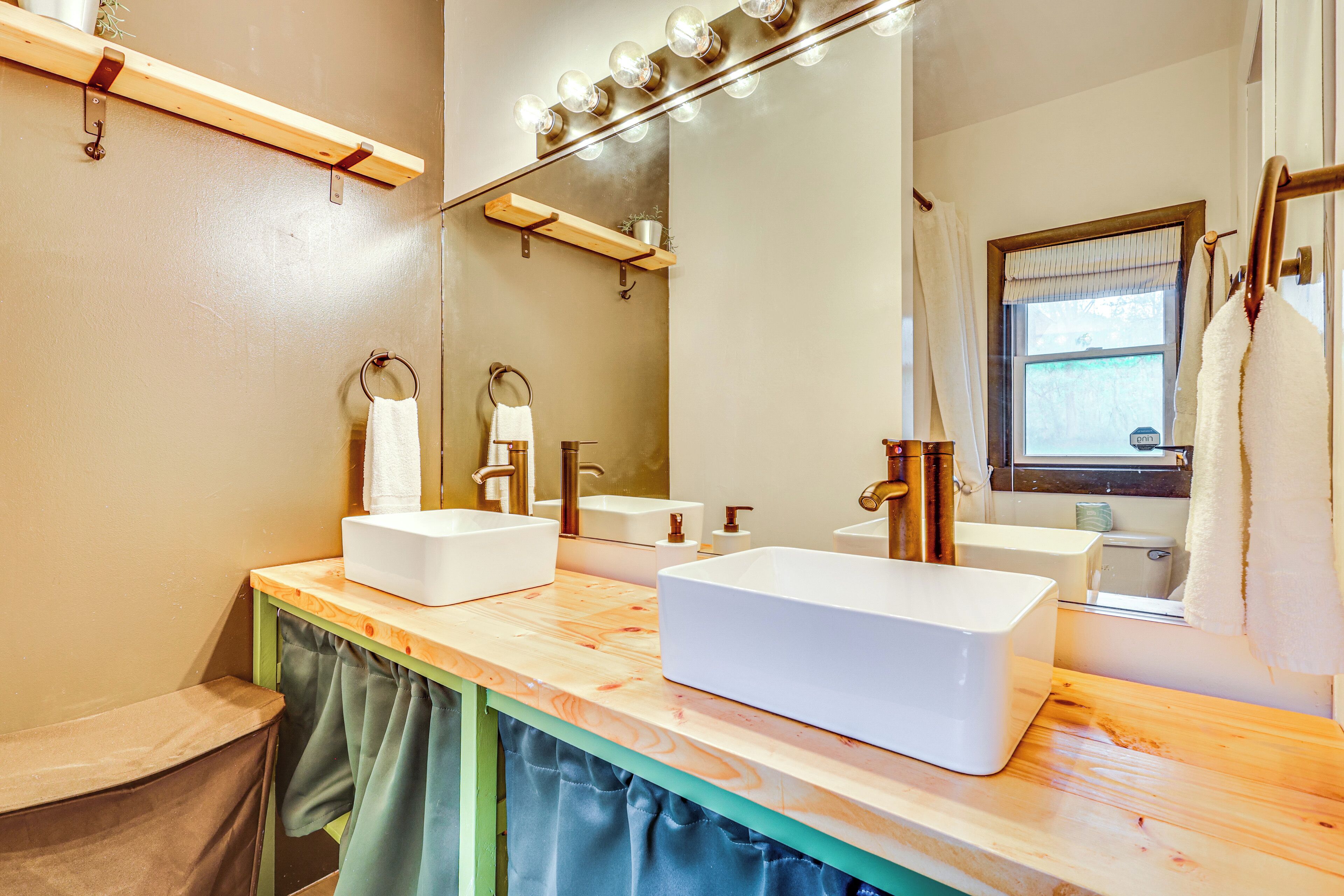 Un baño con dos lavamanos, un espejo con iluminación y una ventana con cortinas.