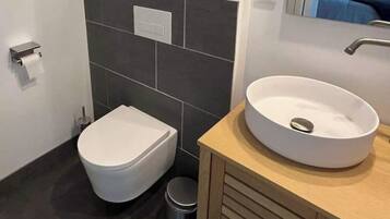 Un baño moderno con inodoro blanco, lavabo redondo y un pequeño cubo de basura.