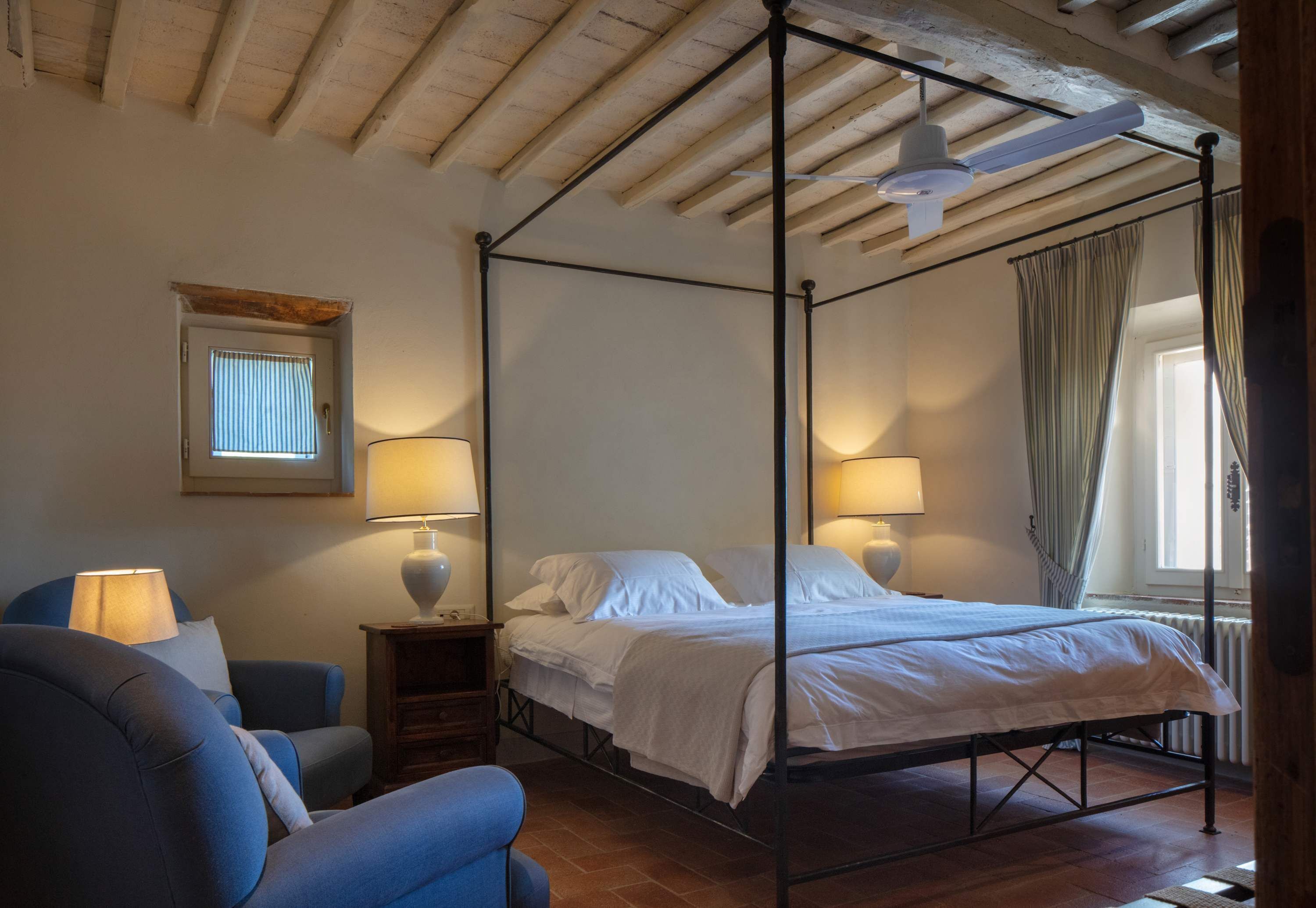 A four-poster bed with white linens, two bedside lamps, a small wooden nightstand, a blue armchair, and a window with curtains.