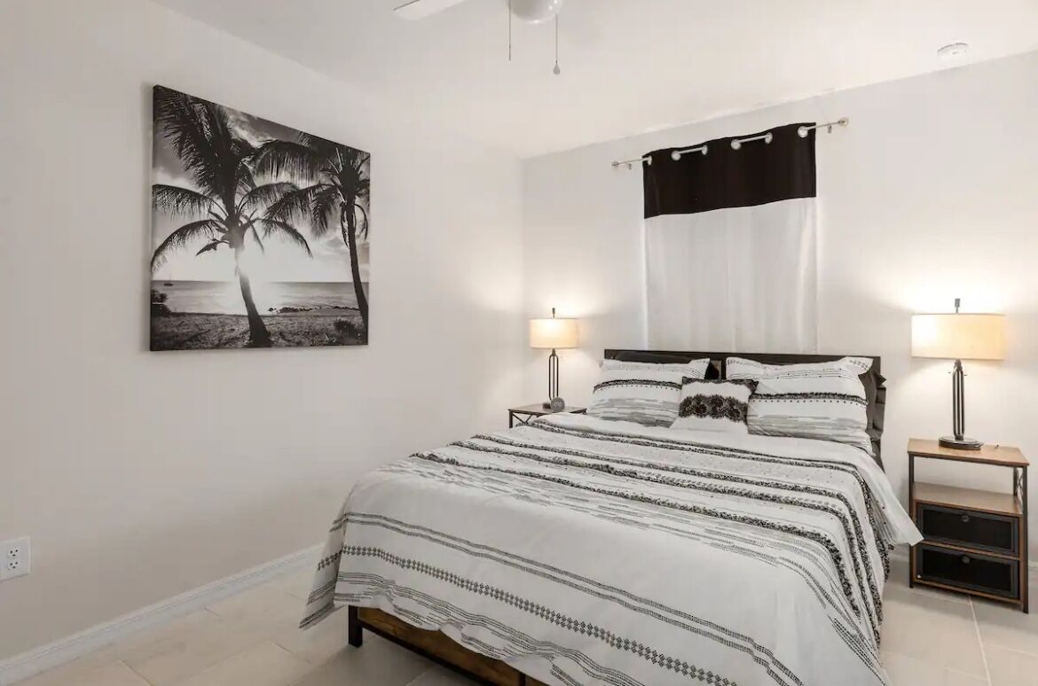 A bedroom with a bed, two bedside tables with lamps, a wall-mounted curtain, and a painting of palm trees.