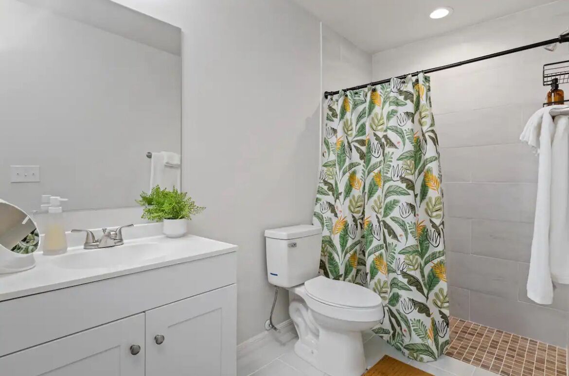Un baño moderno con lavamanos, inodoro y cortina de ducha con motivos de hojas tropicales.