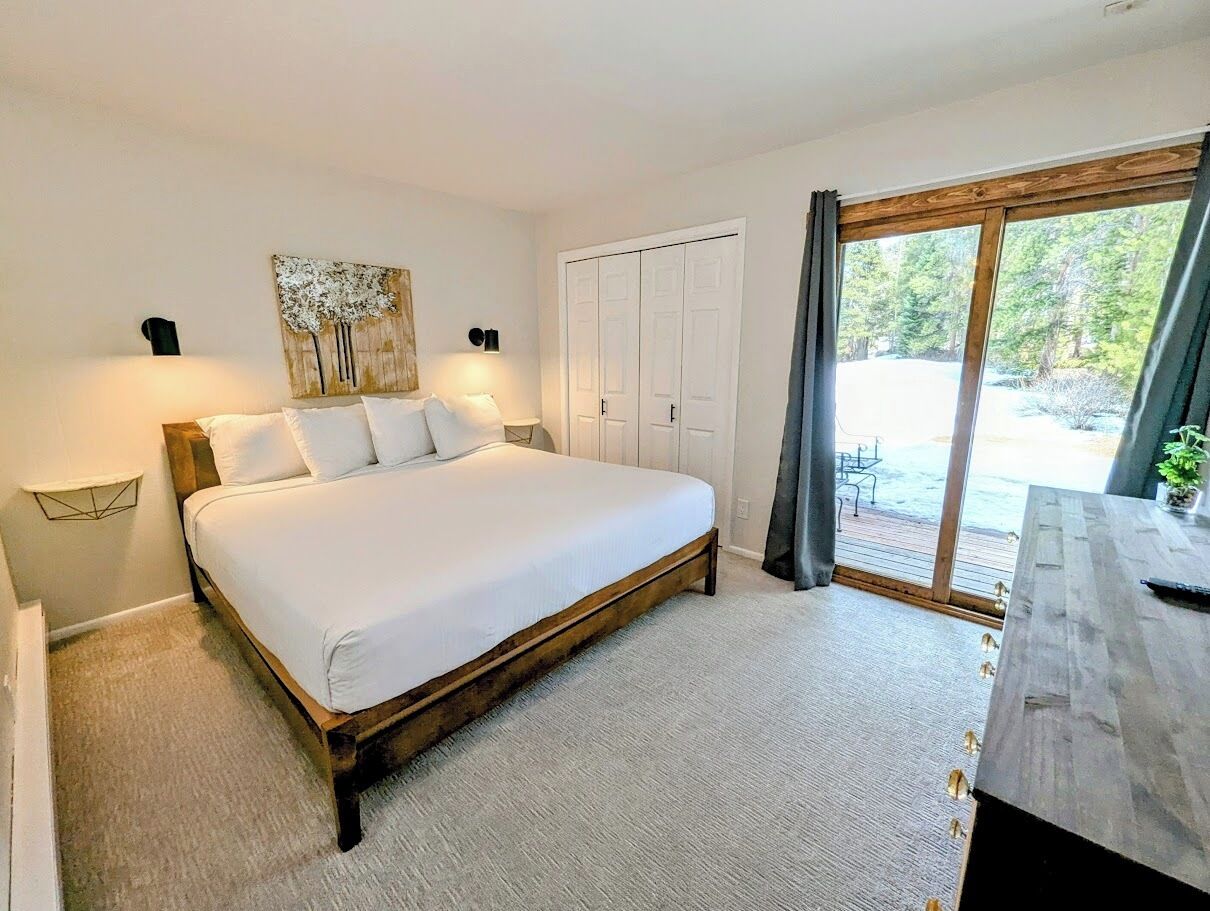 A bedroom with a large bed, a wooden headboard, a nightstand with a lamp, a wardrobe, and a view of the outdoors through a glass door.