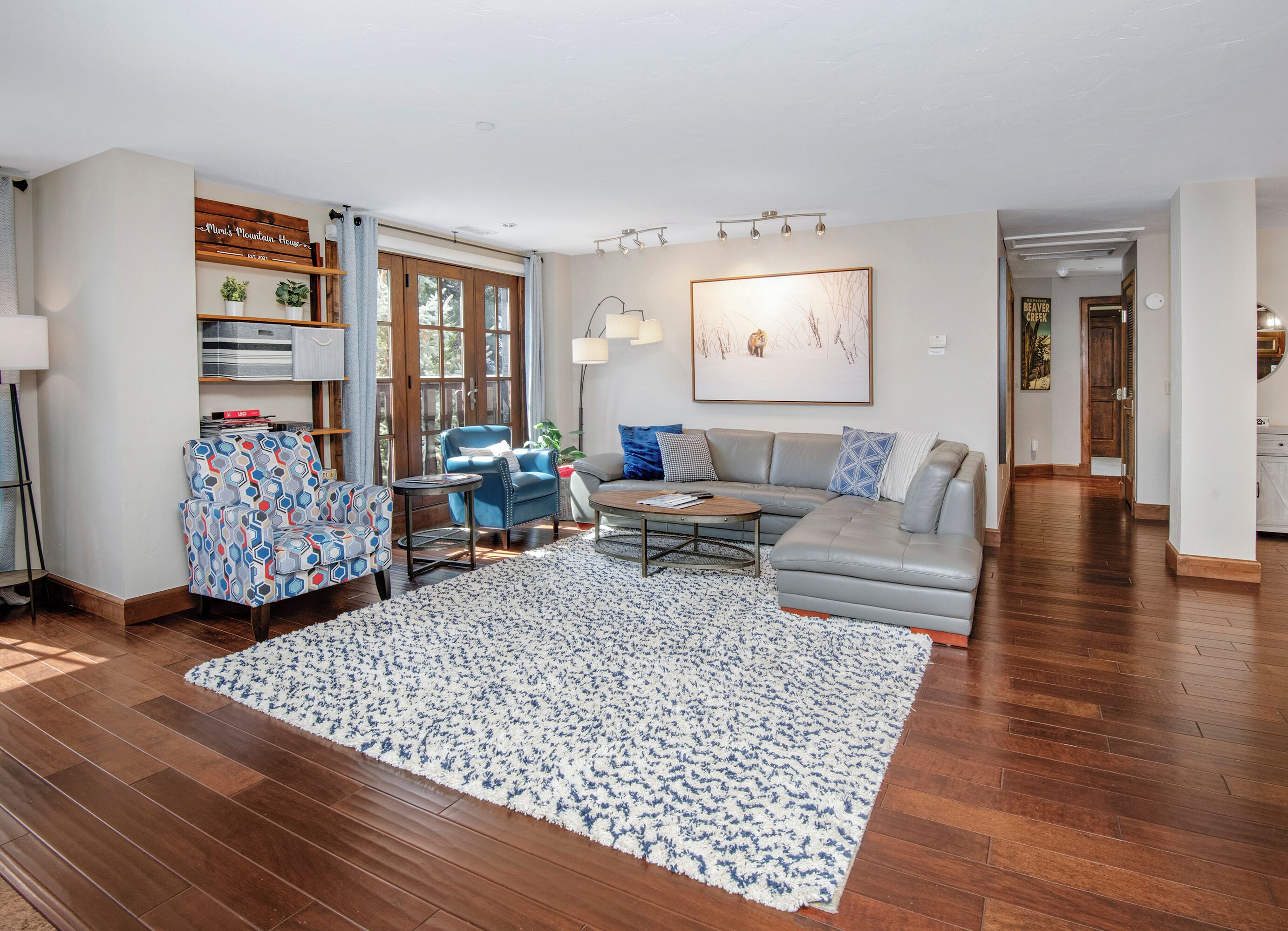 A living room with a gray sofa, blue armchair, and a coffee table. There is a wooden floor, a patterned rug, and a bookshelf with plants.