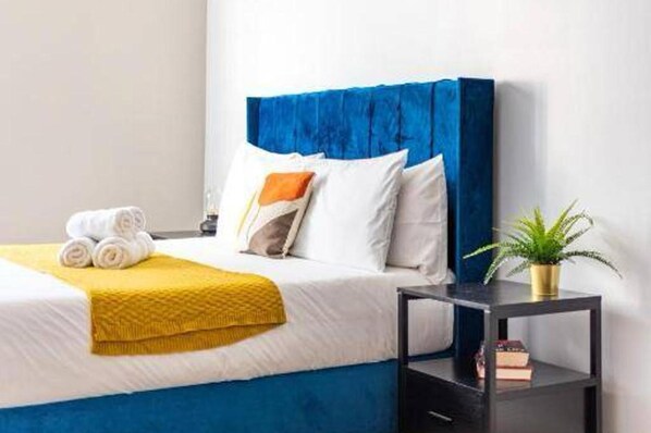 Un lit bien fait, avec une literie blanche et bleue, un plaid jaune et une tête de lit bleue. On y trouve une petite table de chevet avec une plante et quelques livres.