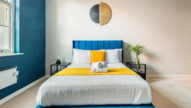 Ein ordentlich eingerichtetes Schlafzimmer mit blauem Kopfteil, einem Bett mit weißer und gelber Bettwäsche, einem Nachttisch mit Lampe und einer runden Wanddekoration.