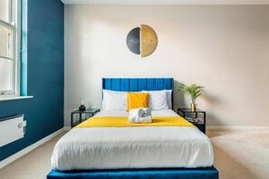 Une chambre à coucher bien rangée, avec une tête de lit bleue, un lit recouvert d’une couette blanche et jaune, une table de chevet avec une lampe et un élément décoratif circulaire fixé au mur.