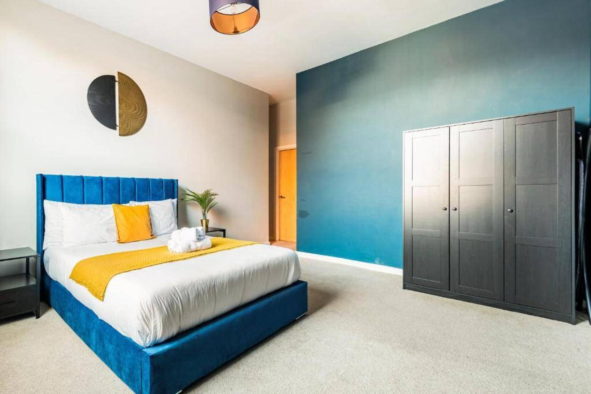 A bedroom with a blue headboard, a bed with white and yellow bedding, a grey wardrobe, and a wall-mounted light fixture.