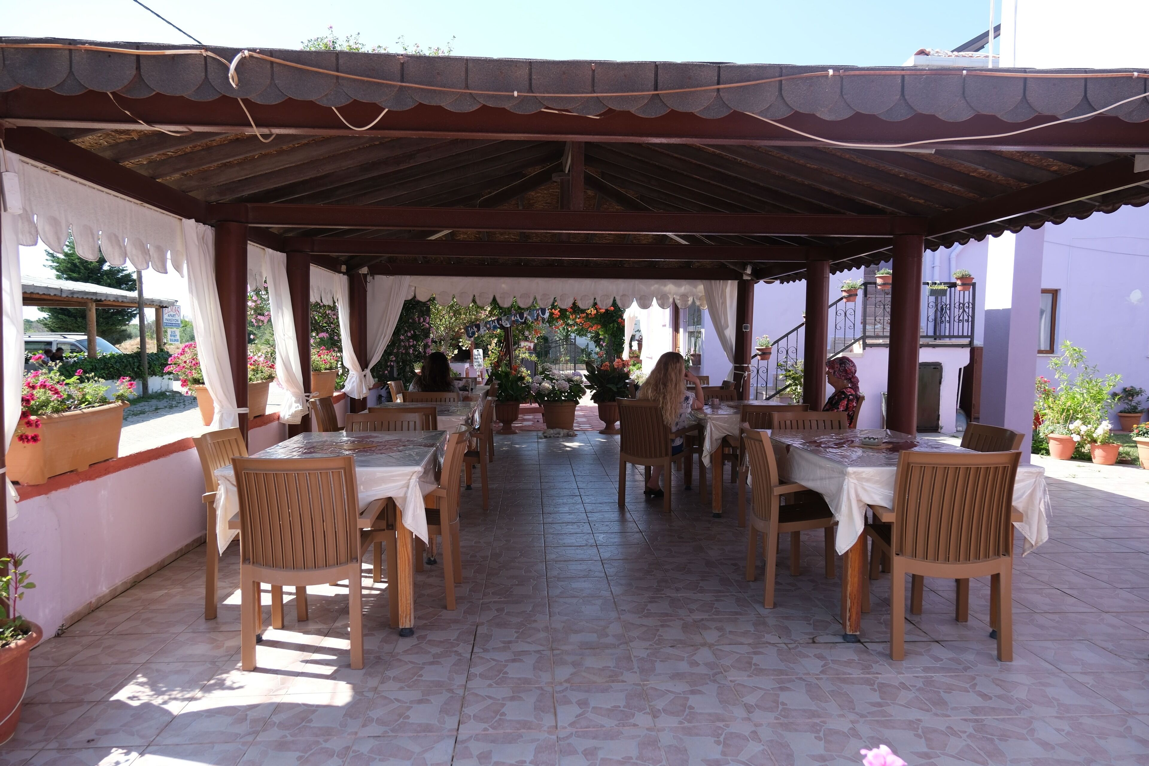 Un patio al aire libre con mesas y sillas de madera, cubierto por un techo de metal, rodeado de plantas y flores en macetas.