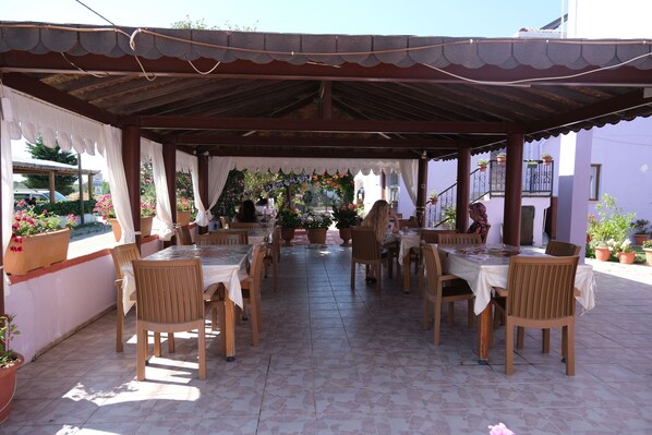 Een terras buiten met houten tafels en stoelen, overdekt met een metalen dak en omringd door potten met planten en bloemen.